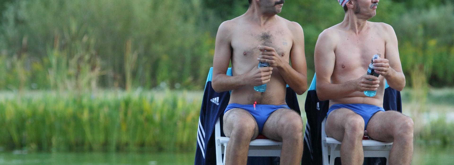 deux hommes en slip et bonnet de bain assis près d'une piscine gonflable regardent vers la gauche