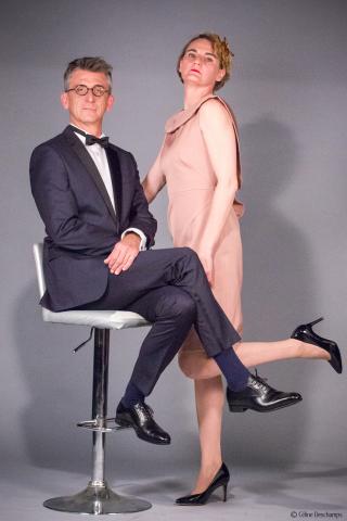 un homme en costume assis sur un taboret haut et une femme en robe rose leve le pied debout